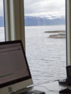 Kontorutsikt fra et av våre sjøanlegg. Vi ser pc-skjerm i forgrunnen, og utsikt til sjøen, merdene og fjellene i bakgrunnen