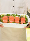 A person holds a serving dish with salmon pieces on vegetables
